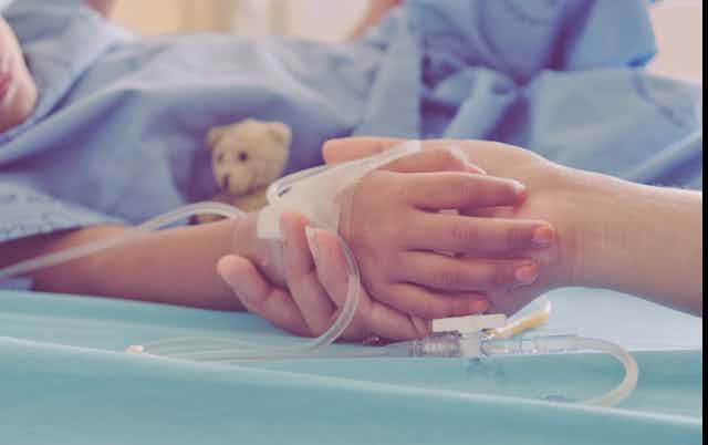An adult's hand holds a child's hand in a hospital setting.
