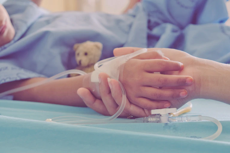 An adult's hand holds a child's hand in a hospital setting.