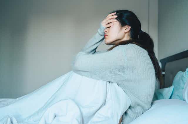 A sick and stressed woman with COVID-19 sits alone in her bed, her hand on her forehead.