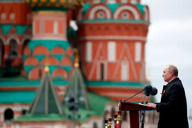 A balding man stands at a podium with microphones in front of him, colourful buildings in the background