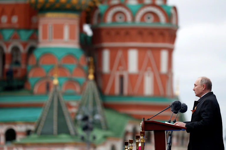 A balding man stands at a podium with microphones in front of him, colourful buildings in the background