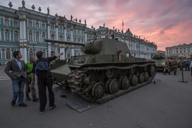 People look at a Second World War-era tank as the sun rises in a city square.