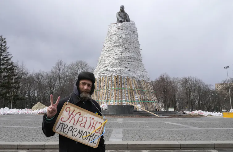 A bearded man holds a poster reading 'Ukraine will win' against the background of a monument of a Ukrainian hero covered with sandbags to protect it from Russian shelling.