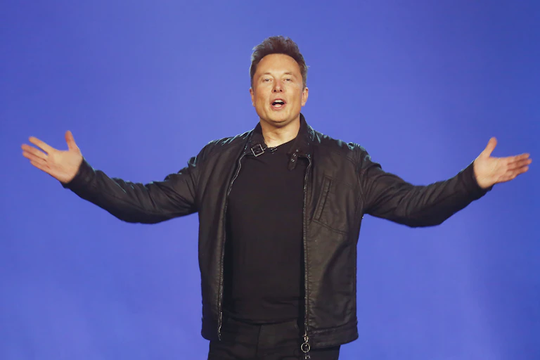 A white man wearing black clothing stretches both arms out wide with open palms as he appears to be speaking with an open mouth, on a blue backgorund