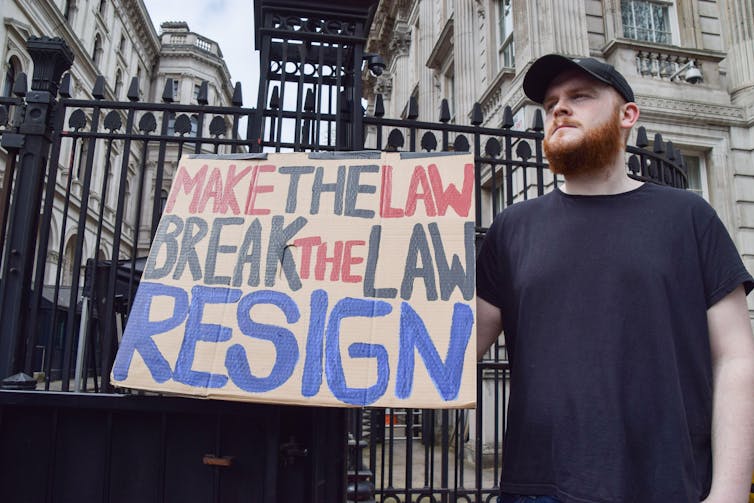 A protestor holds up a sign reading 'make the law, break the law, resign' outside the gates of Downing Street.