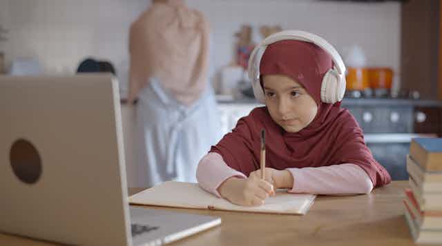 a young girl wearing the hijab takes notes as she looks at a laptop and wears headphones