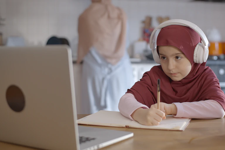 a young girl wearing the hijab takes notes as she looks at a laptop and wears headphones
