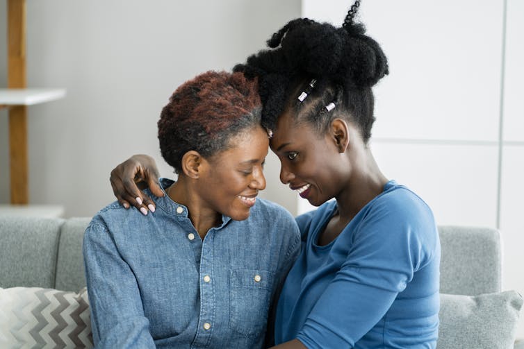 A young black woman embraces her mother while seated on a grey couch.