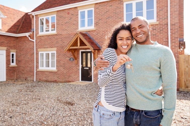 A young couple stands outside a brick house holding up a set of keys.