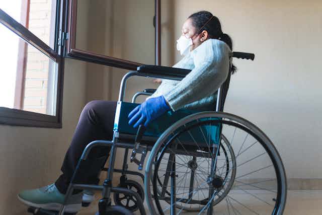 A woman sitting in a wheelchair looking out of an open window. She is wearing a face mask and rubber gloves.