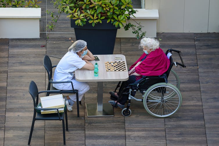 View from above of a nurse and elderly woman in a wheelchair, both wearing face masks, sitting at a table in a courtyard playing checkers.