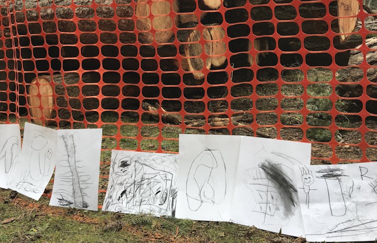 A stack of cut trees seen behind a mesh fence that is lined with children's drawings.