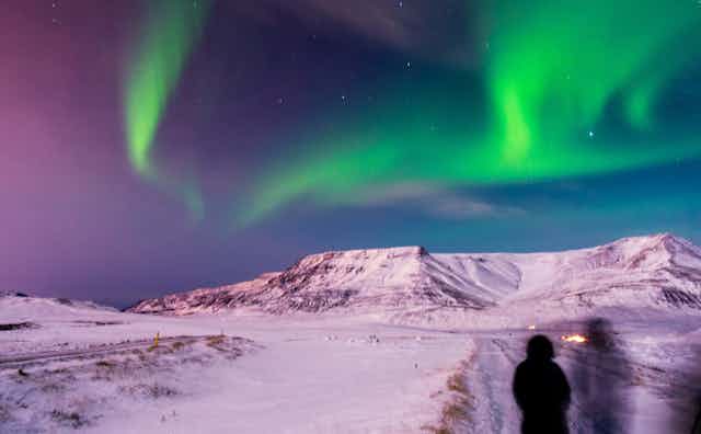 A person stands with a snowy landscape and mountains ahead and the Northern Lights in the sky overhead.