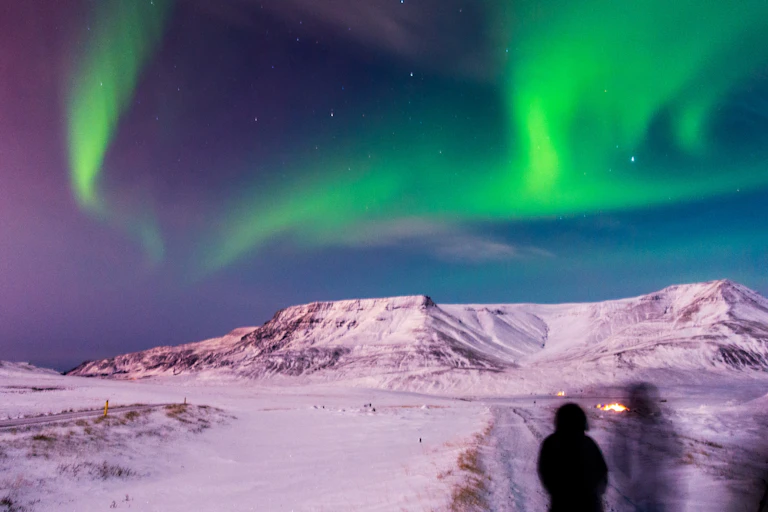 A person stands with a snowy landscape and mountains ahead and the Northern Lights in the sky overhead.