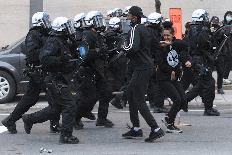A swarm of police in riot gear encircle two young people dressed in black.
