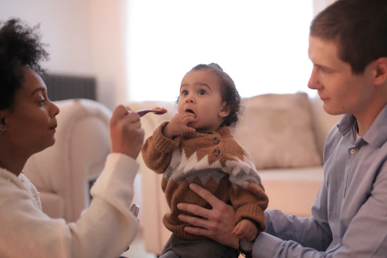 Two parents are seen holding a spoon to feed a baby.