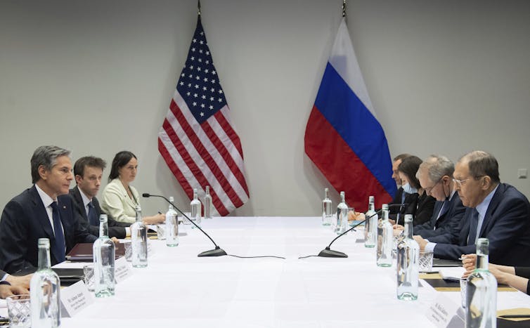 Six people sitting around a table. Blinken and Lavrov are making eye contact from opposite sides of the table. The U.S. and Russian flags are in the background.