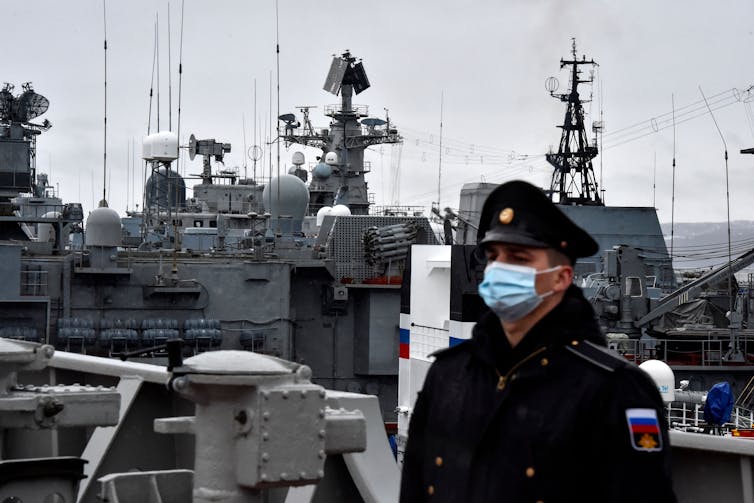 A servicemember in a coat and military hat stands with military ships behind him.