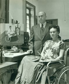 A man and a woman in a wheelchair pose together next to a painting.