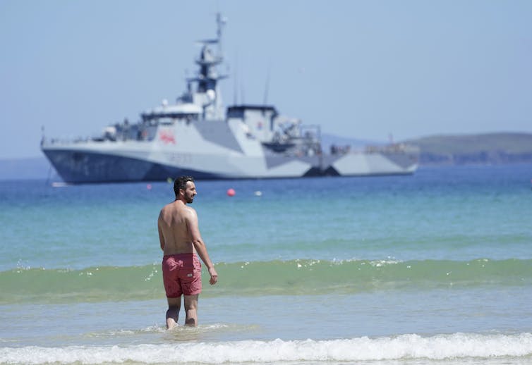 A man in a red bathing suit stands in the surf with a large patrol ship in the background.