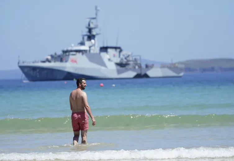 A man in a red bathing suit stands in the surf with a large patrol ship in the background.