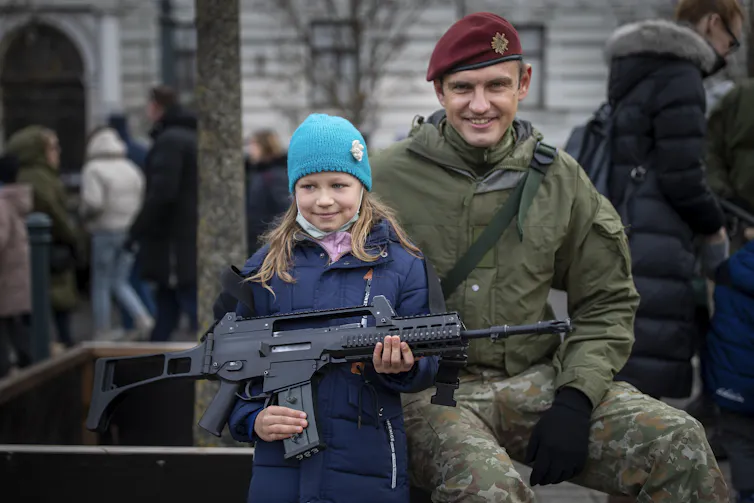 A girl in a bright blue knit hat holds a military rifle as a uniformed soldier in a dark red beret smiles sitting next to her.