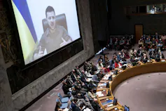 Zalensky is seen speaking on a video screen with a blue and yellow flag behind him.