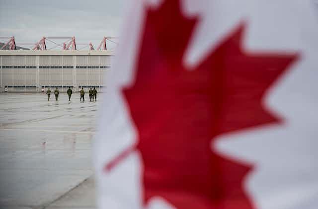 Uniformed Canadian military personnel walk on a tarmac with the Canadian flag in the foreground.
