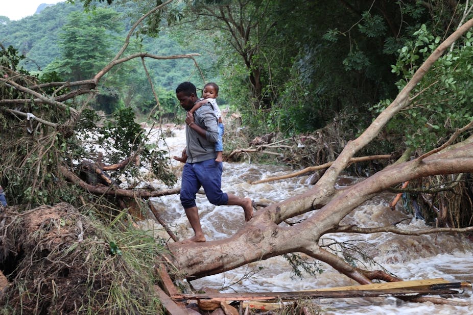Man holding a baby on his back using a tree to cross a gushing river.