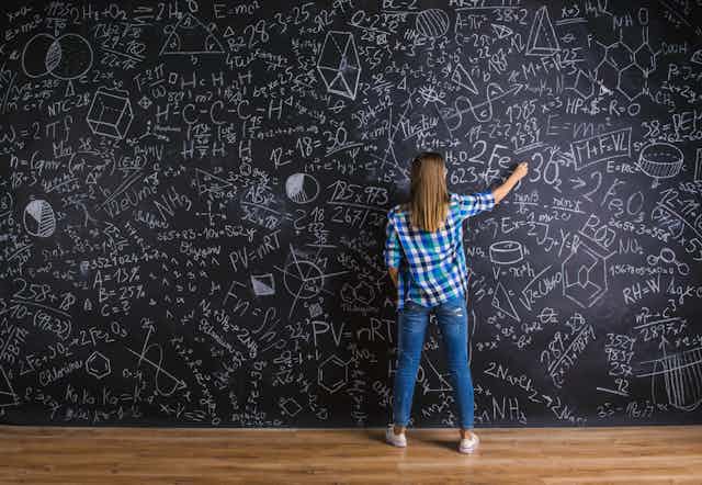 Mujer de espaldas escribiendo en una pizarra llena de ecuaciones.