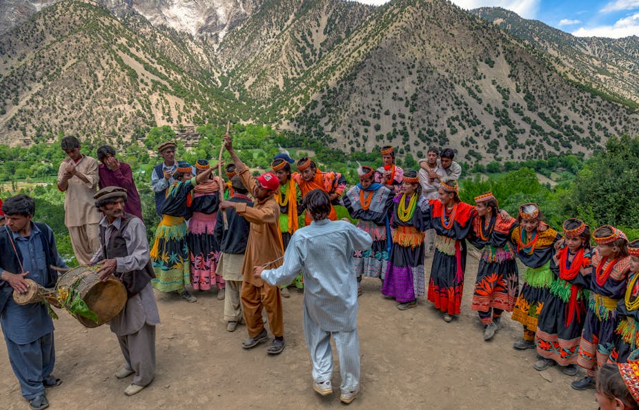 Men with musical instruments with dancers in colourful clothes behind and mountains in the background.
