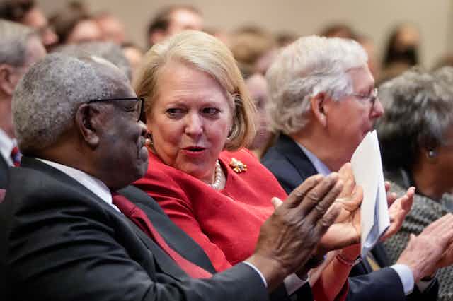 In the audience at a gathering, a man with gray hair in a dark suit and red tie sits next to a blond woman in a red suit who is talking to him.