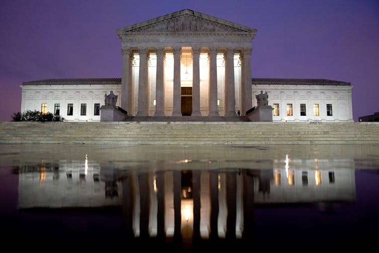 A tall white stone building with eight columns and two wings, overlooking marble steps and a puddle in which it is reflected.