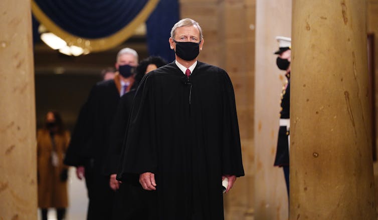 A gray-haired man with a black mask and wearing a black robe walks down a corridor.