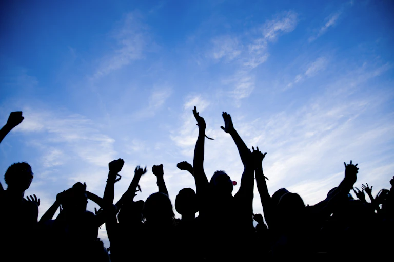 A row of people joyously raise their hands - the people are shadowed against a blue sky