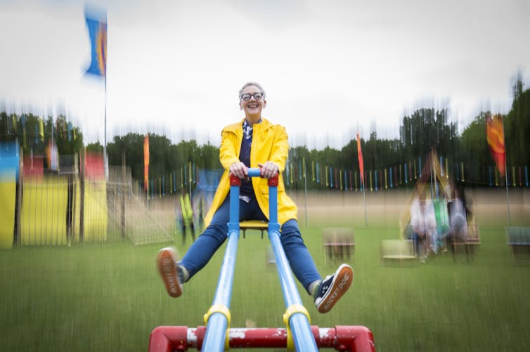 A woman in a bright yellow jacket and blue pants sits on a seesaw, smiling, and facing the camera behind a blurred b background.