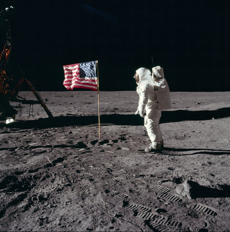 Buzz Aldrin in a spacesuit on the surface of the Moon next to the US flag.