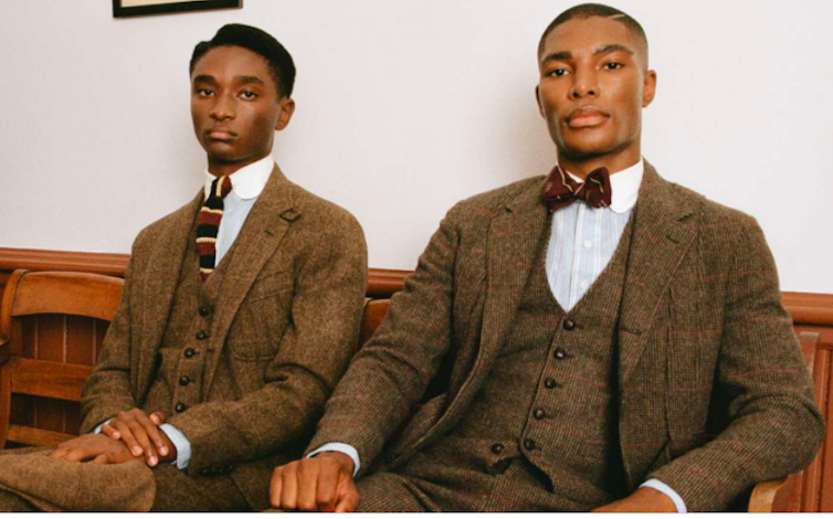 Two black men are wearing business suits and ties while sitting on a bench.
