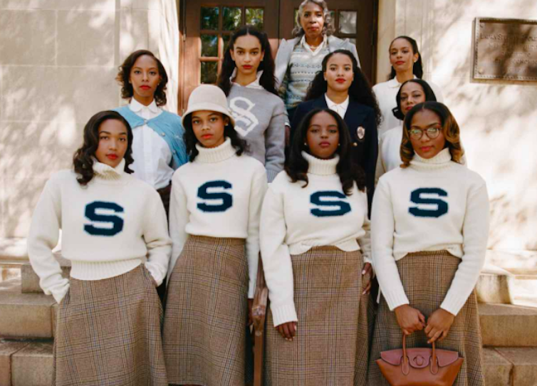 Some of the black women standing in front of a building are wearing white turtleneck sweaters with the letter 'S'.