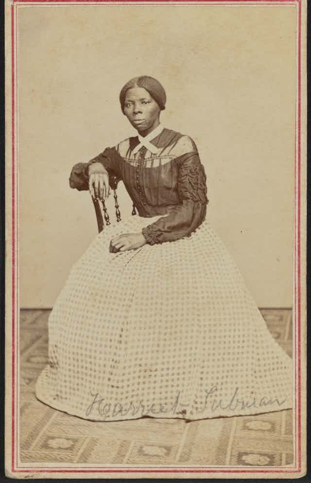 A black woman dressed in a black and white gown is sitting on a chair. 