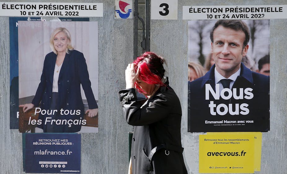 A woman with red hair scratches her head as she walks past posters of Emmanuel Macron and Marine Le Pen.