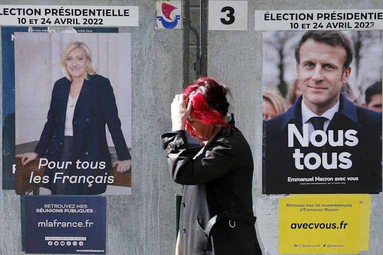 A woman with red hair scratches her head as she walks past posters of Emmanuel Macron and Marine Le Pen.