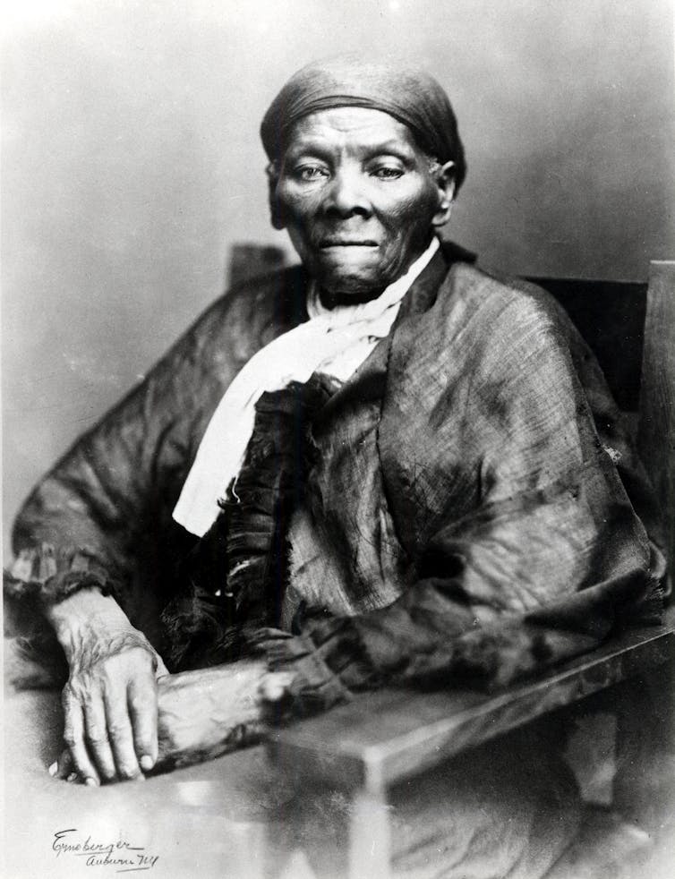 An elderly Black woman holds her hands as she sits in a chair and poses for a portrait.