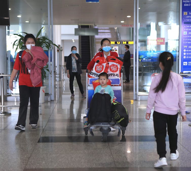 A mother wearing a mask pushes a young child on a luggage cart at an airport as a young girl walks towards them.