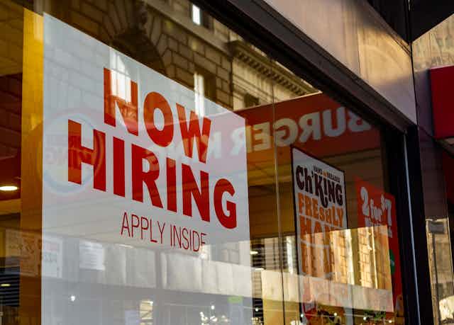 A 'Now Hiring' sign hanging in a shop window