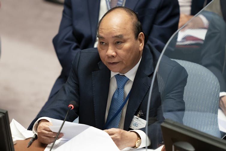 A man speaks into a microphone with papers on the desk in front of him.