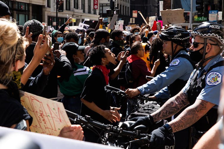 A crowd of people including some holding signs and some shouting, face police officers on bicycles at a large protest.