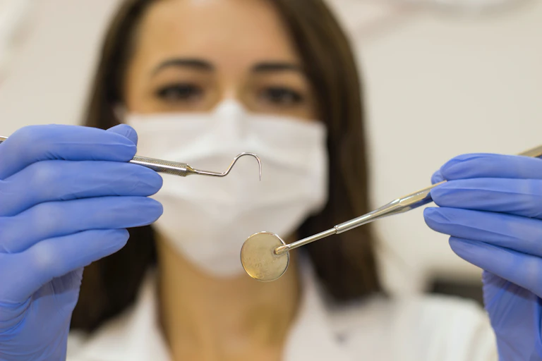 A woman in white coat and face mask out of focus in background with her gloved hands holding dental instruments in the foreground
