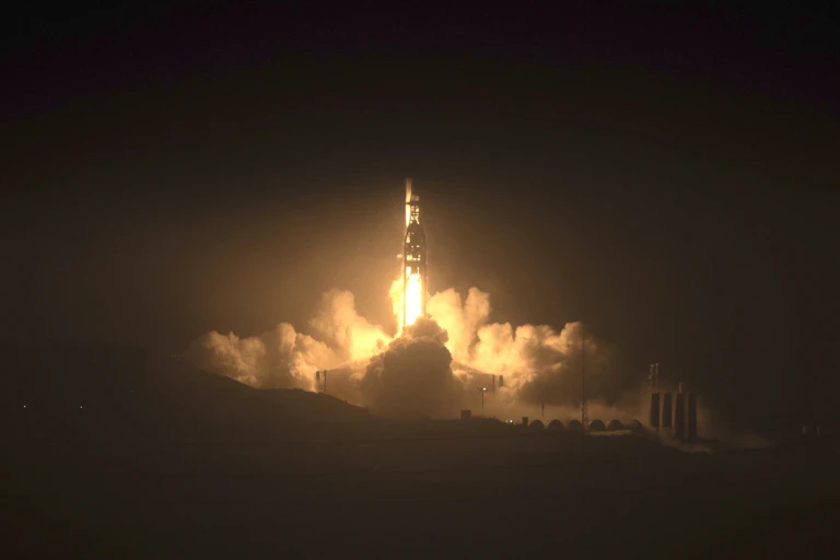 A rocket launches amid smoke and flames against a dark landscape.
