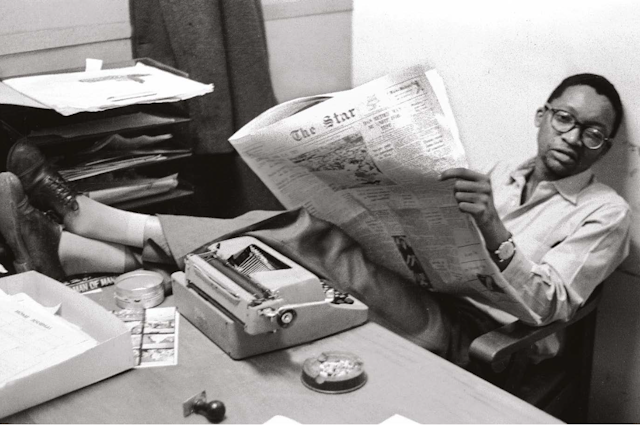 A man in glasses leans back in his chair, a newspaper open in front of him, his feet crossed on the desk next to a typewriter.
